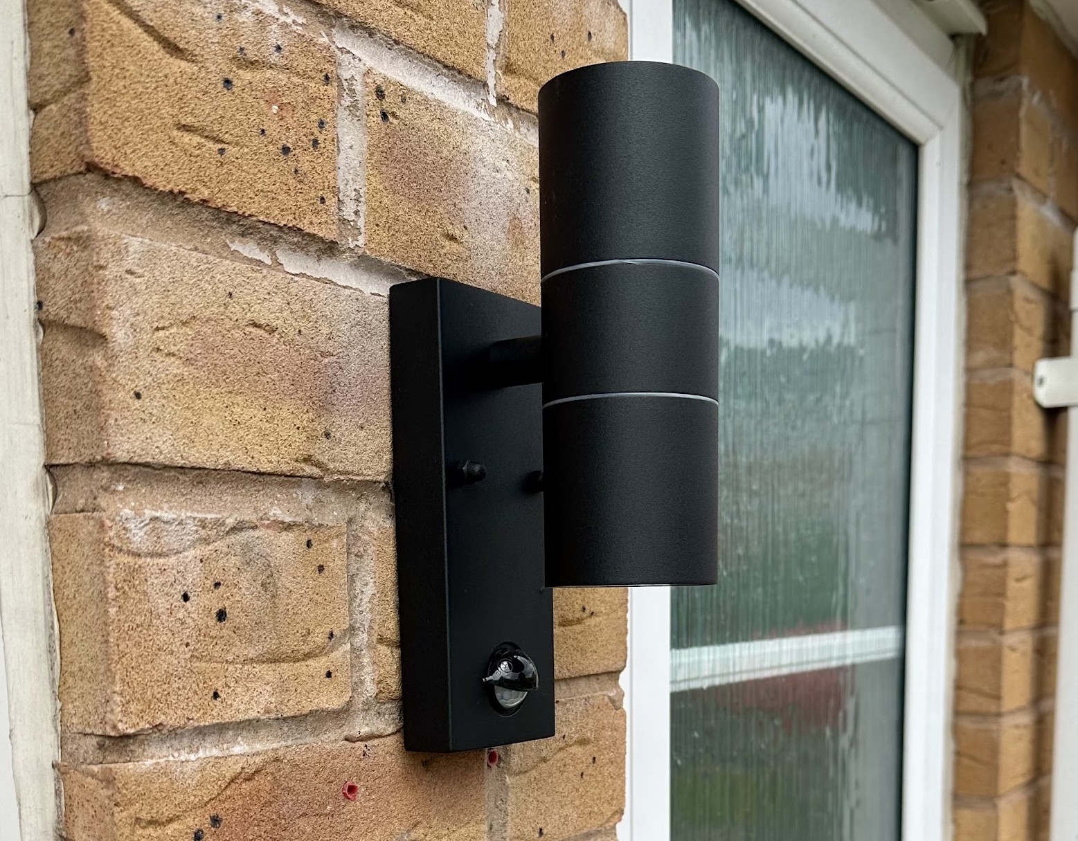 A black up and down outdoor wall light with a sensor, fitted to a brick wall beside a front door