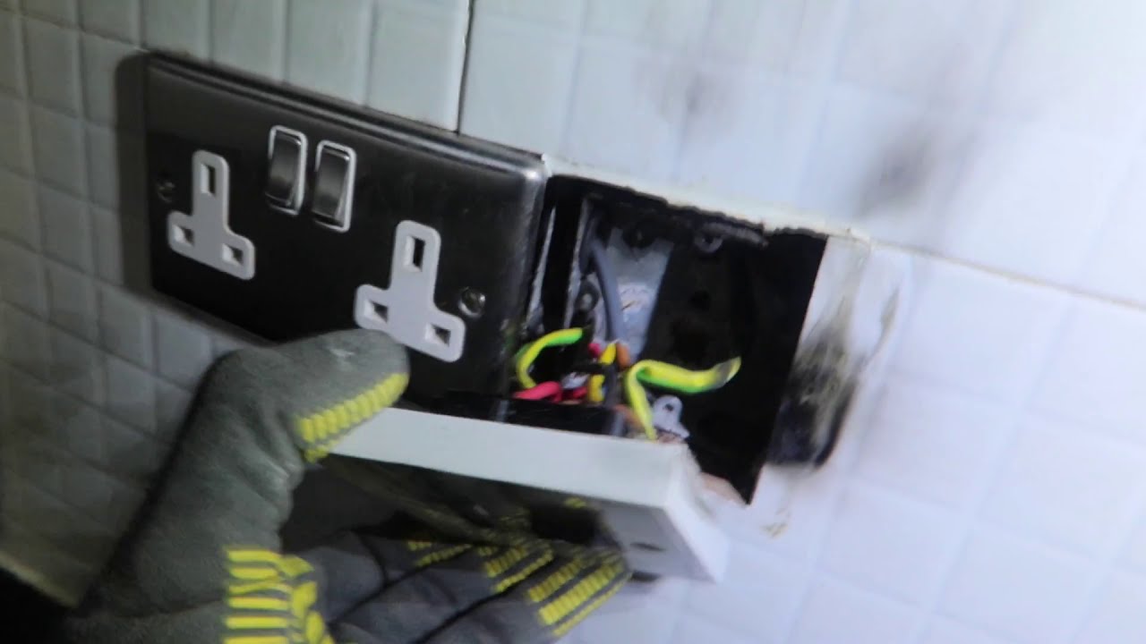 A scorched double socket pulled away from a tiled kitchen wall, held in a gloved hand