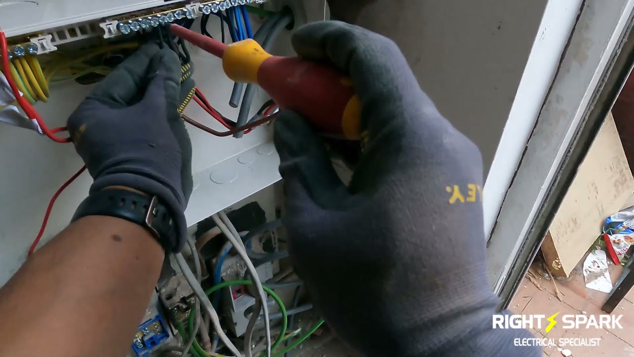 Gloved hands tightening cable terminals inside a consumer unit